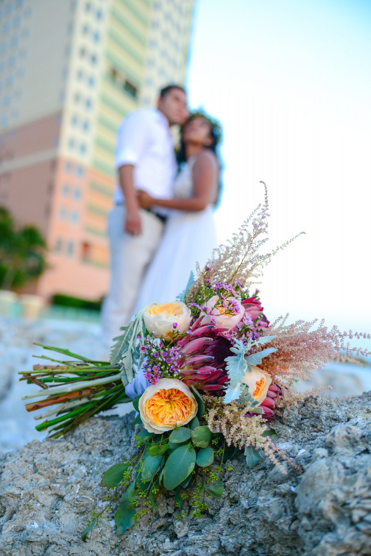 Wedding bouquet in focus on rocks with blurred couple and building in background.