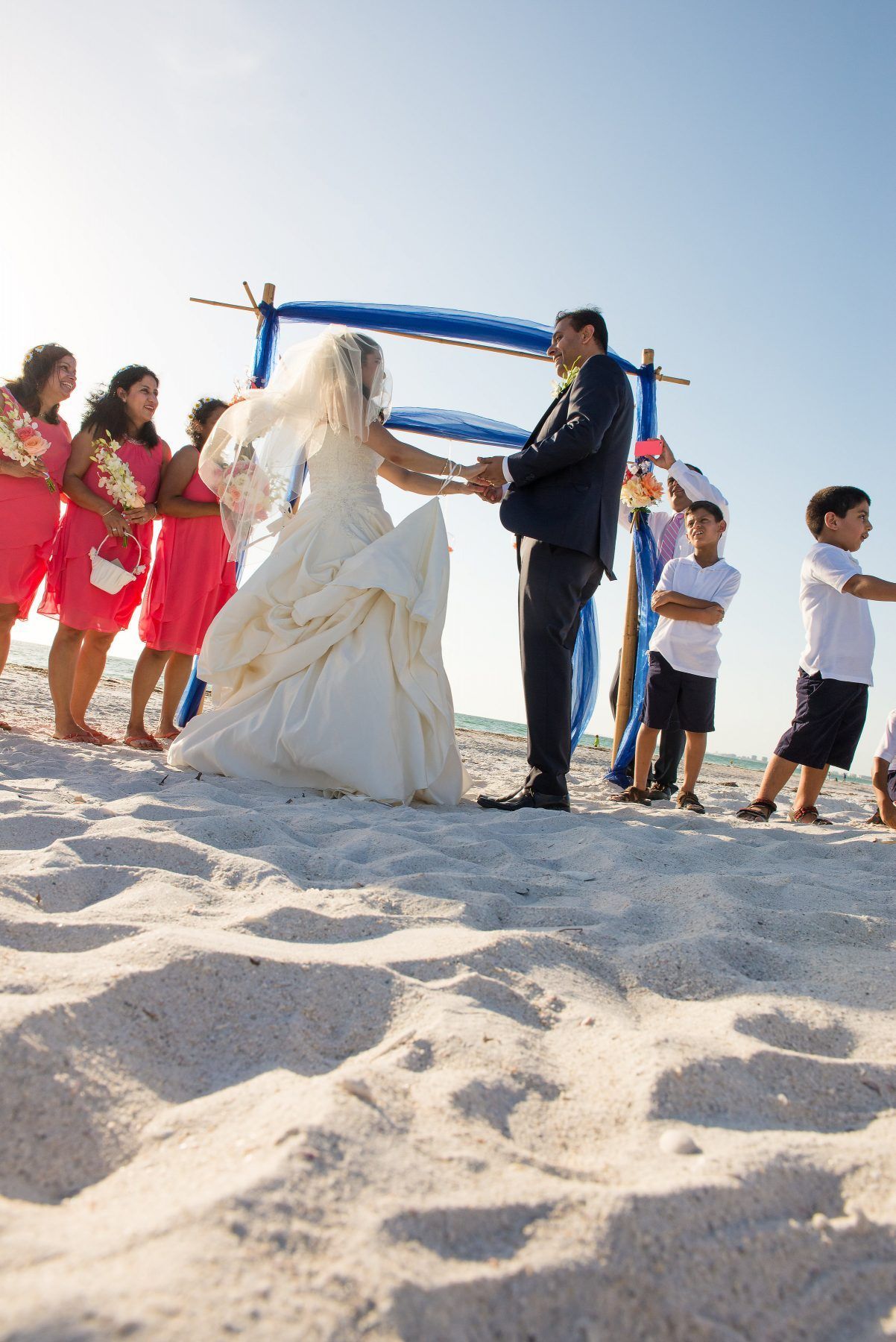 Bride and groom hold hands at beach wedding. Bridesmaids in coral dresses stand near arch.