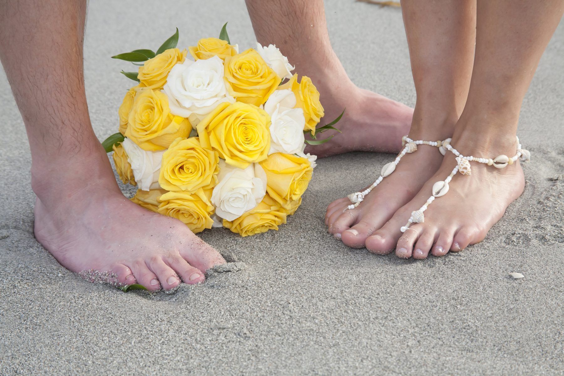 Bare feet stand near a yellow and white rose bouquet on a sandy beach. One person wears a shell anklet.