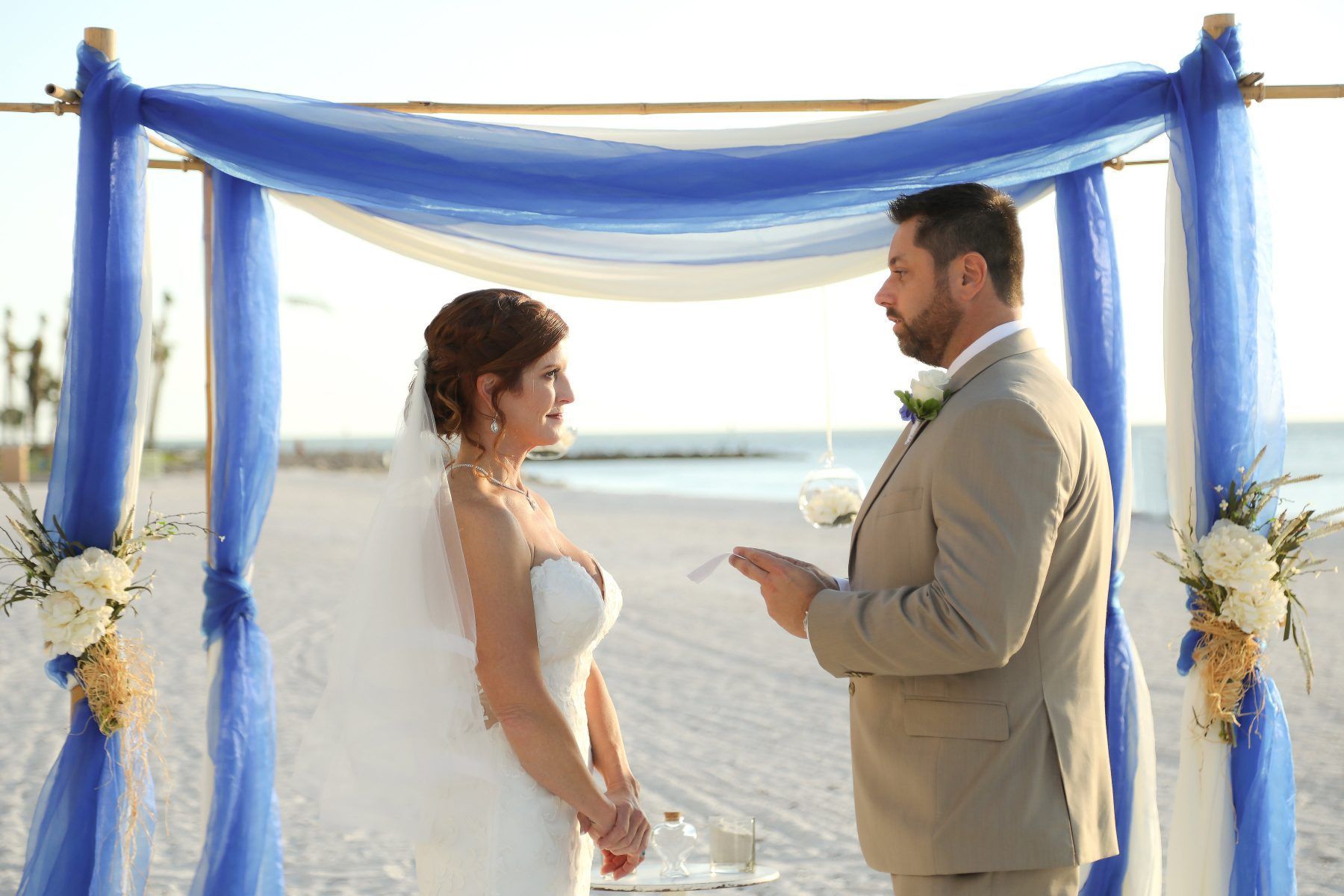 Bride and groom at beach wedding ceremony under blue draped arch.