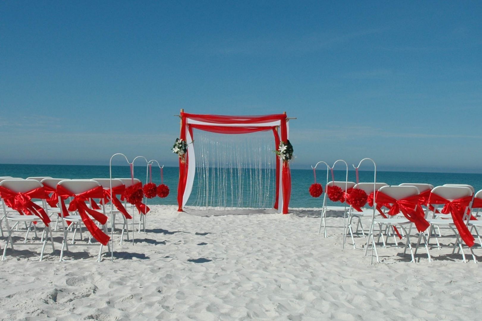 Beach wedding ceremony setup with white chairs and red decorations against a blue sky.