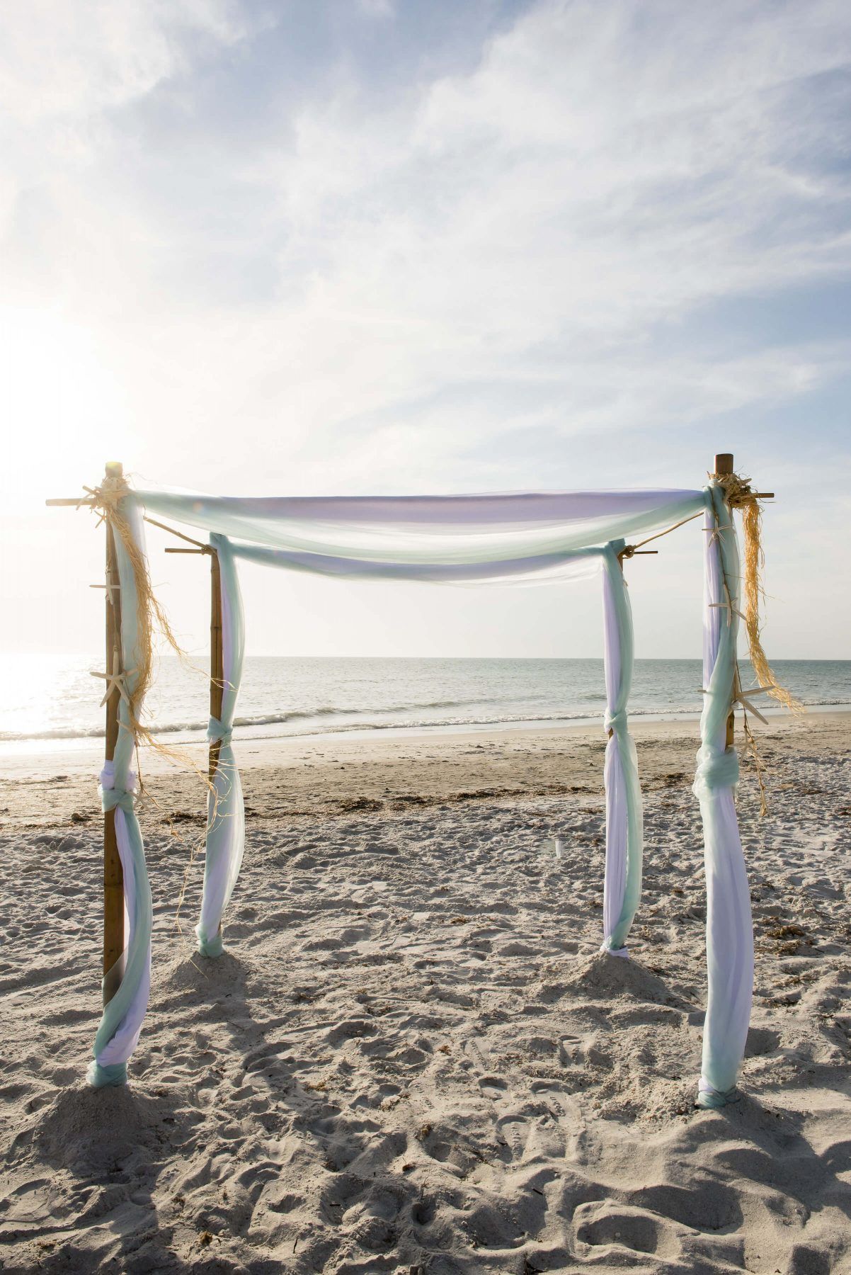 Wedding arch on a sandy beach, draped with pastel fabric. Ocean and bright sky in the background.
