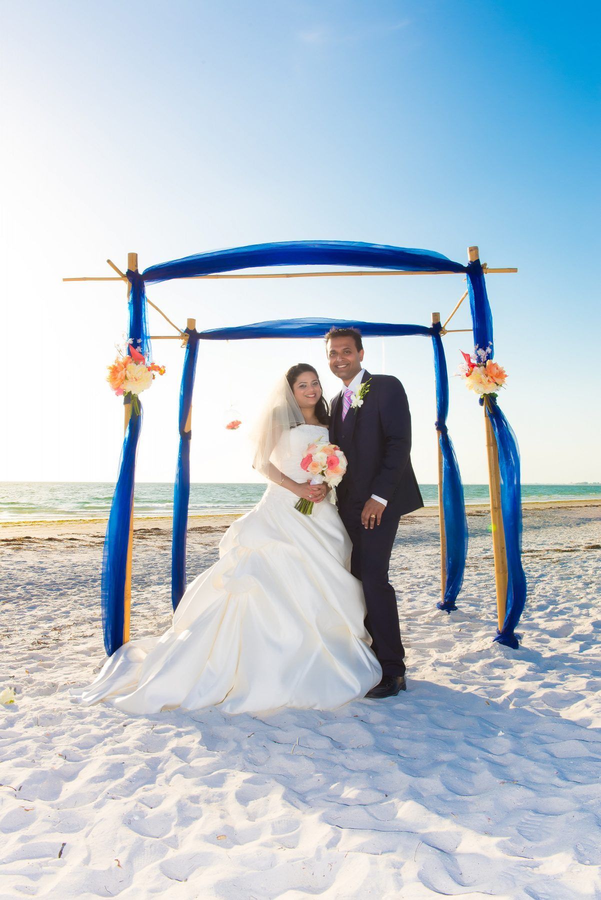 Bride and groom standing on beach under a blue draped arch. Bride in white dress, groom in suit. Blue sky.