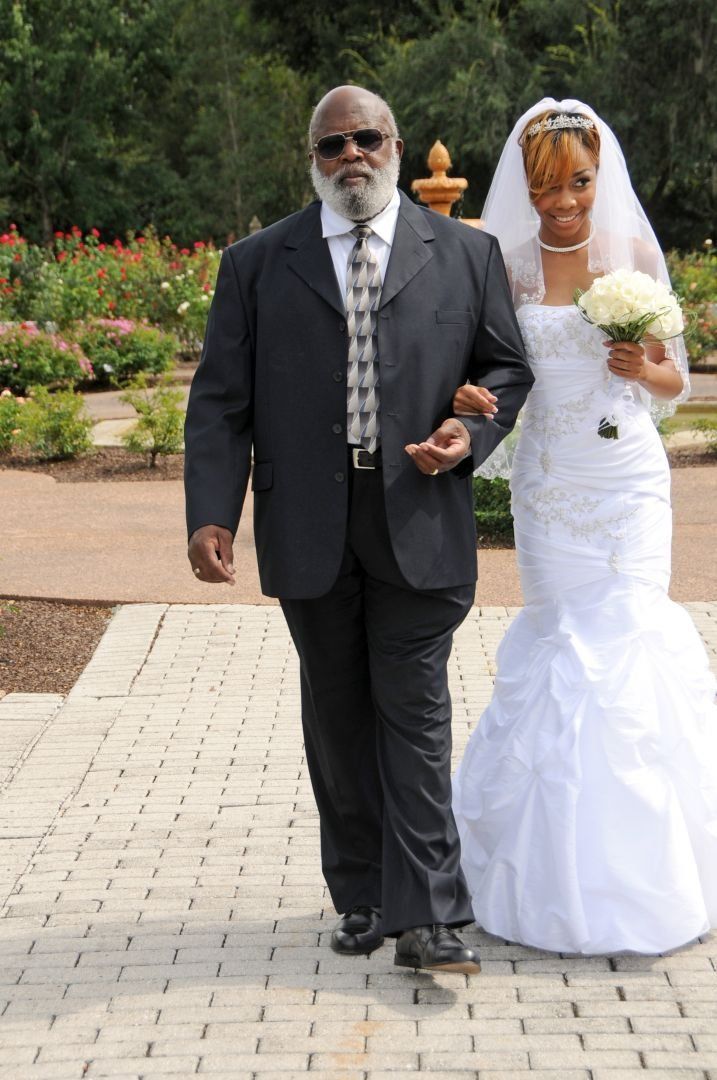 Father escorting bride down a brick path, holding her arm, at an outdoor wedding. She wears a white gown and veil.
