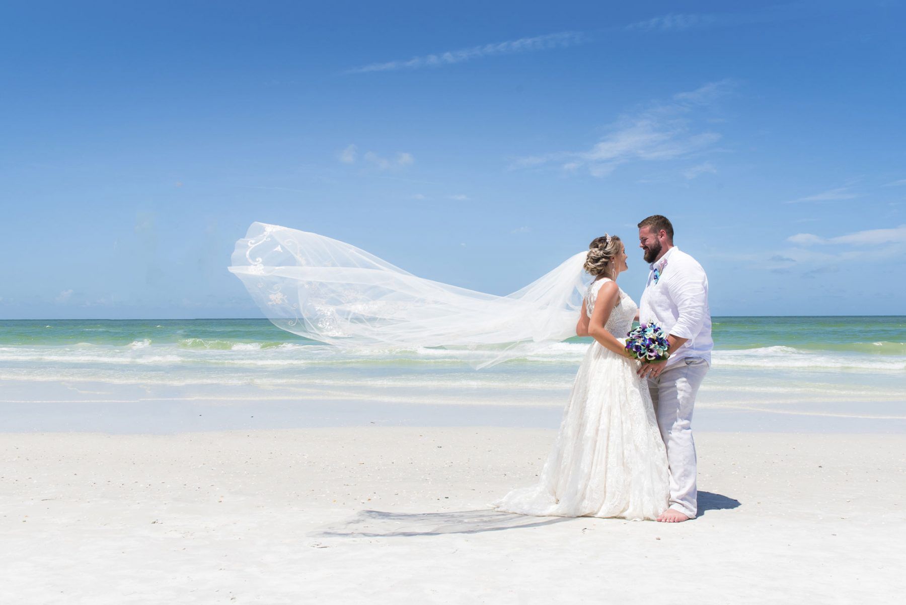 Bride and groom on a beach, veil blowing in the wind, sunny day.