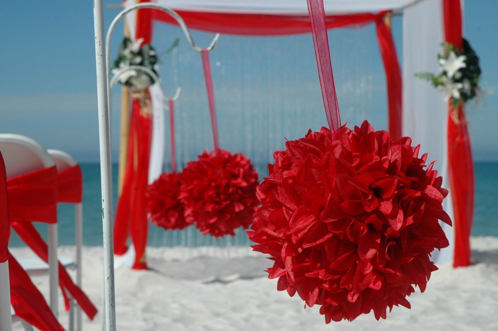 Red floral decorations at a beach wedding ceremony, with white chairs, arch, and ocean background.