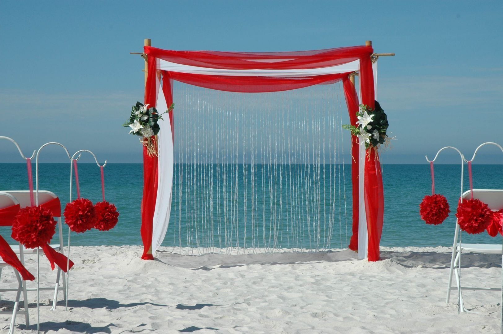 Beach wedding arch with red and white fabric against a blue sky, white sand, and chairs with red decorations.