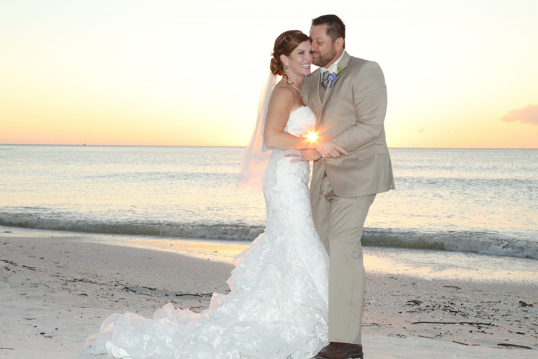 Couple embraces on beach at sunset; bride wears white dress, groom in tan suit.
