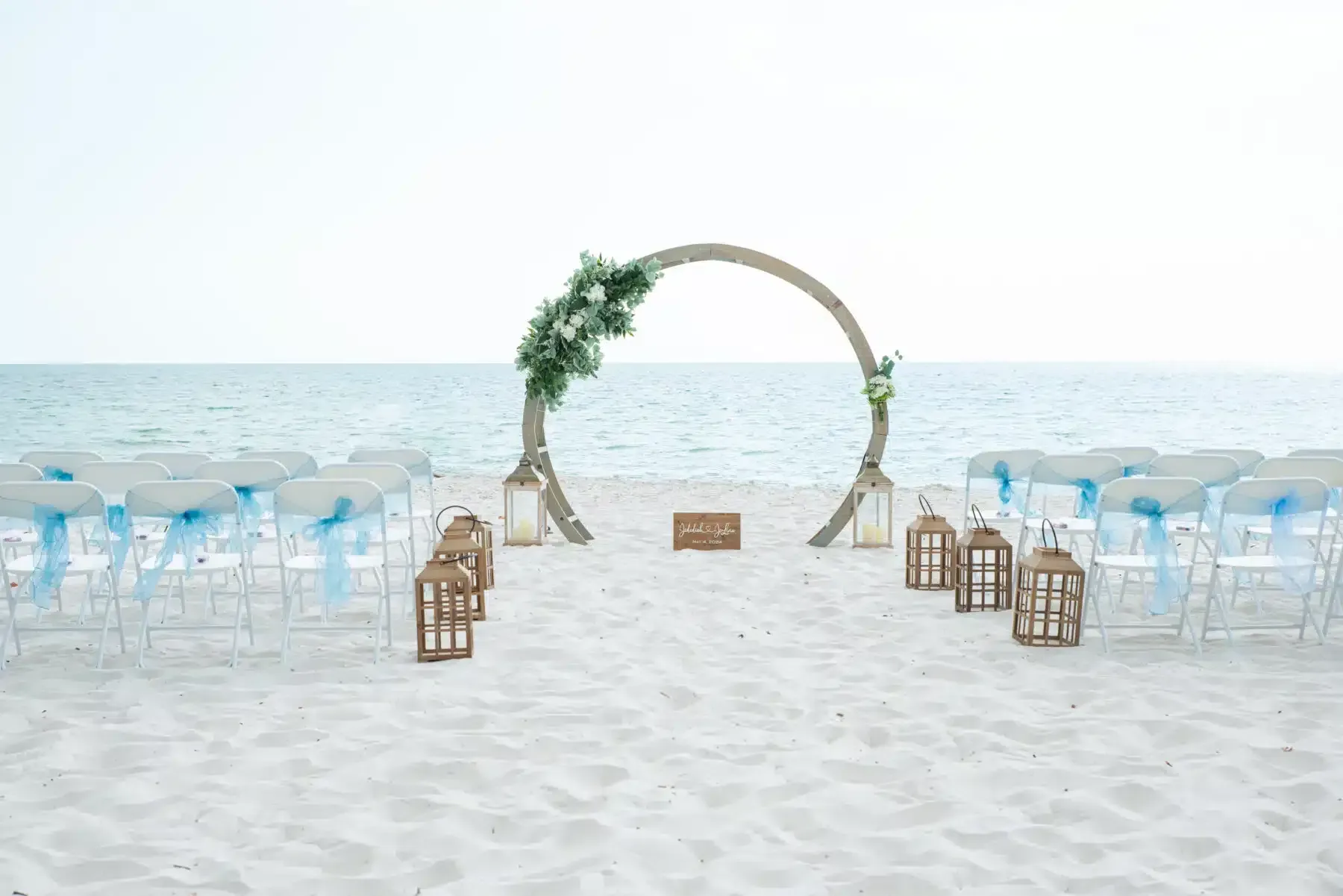 Beach wedding ceremony setup with an arch decorated with greenery and chairs with blue ribbons.
