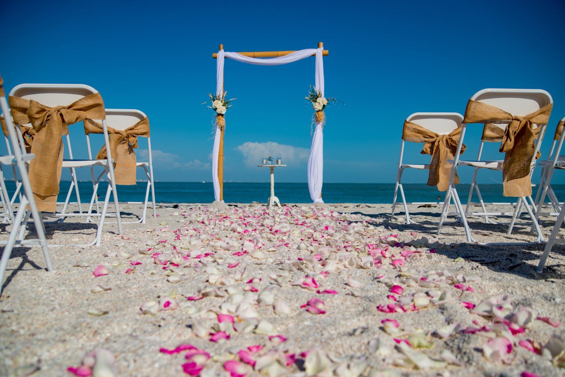 Beach wedding ceremony setup: arch, chairs with burlap bows, and rose petals on the sand, blue sky and ocean in the background.
