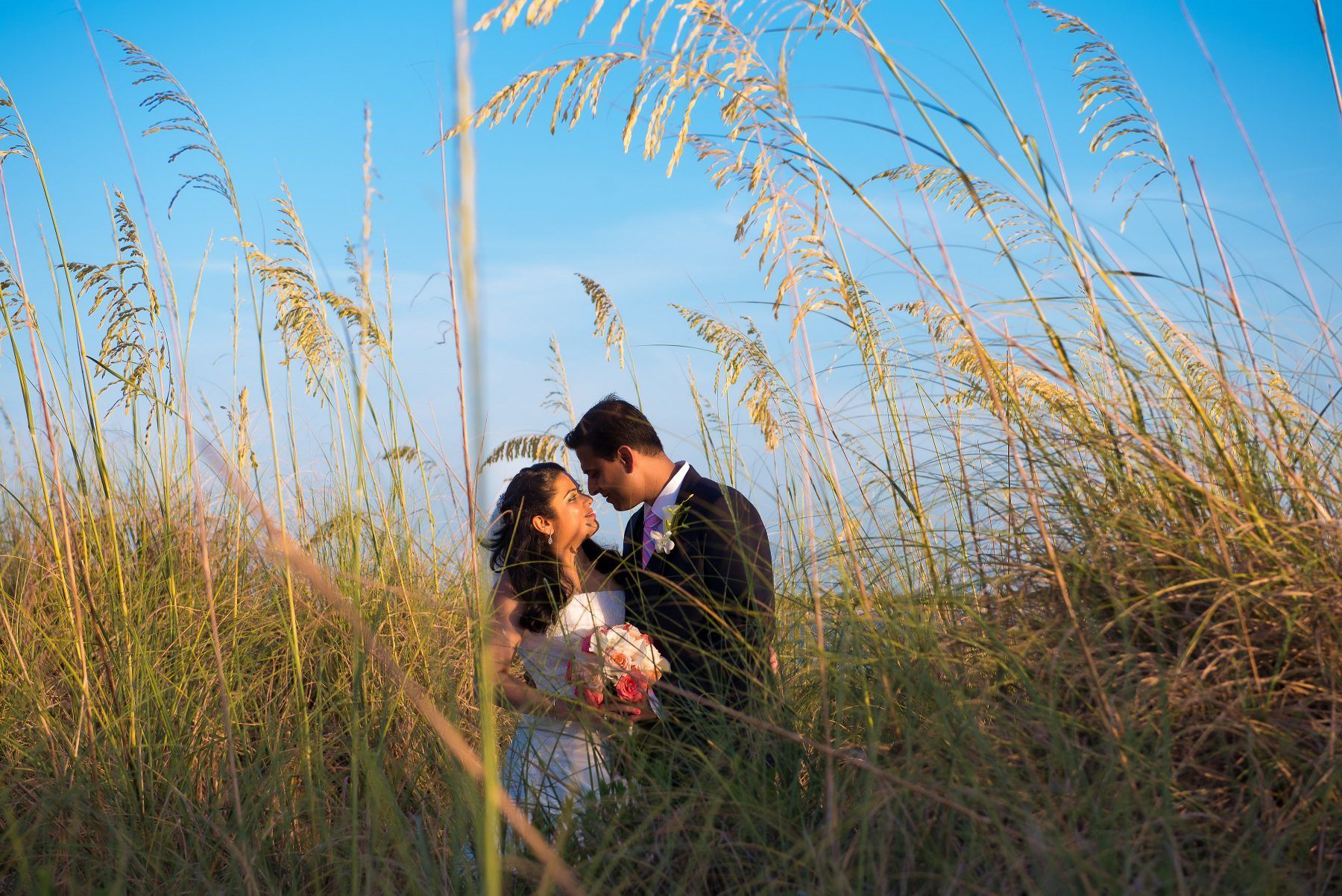 Couple embraces amongst tall grass, blue sky background. Man in suit, woman in white dress, holding flowers.