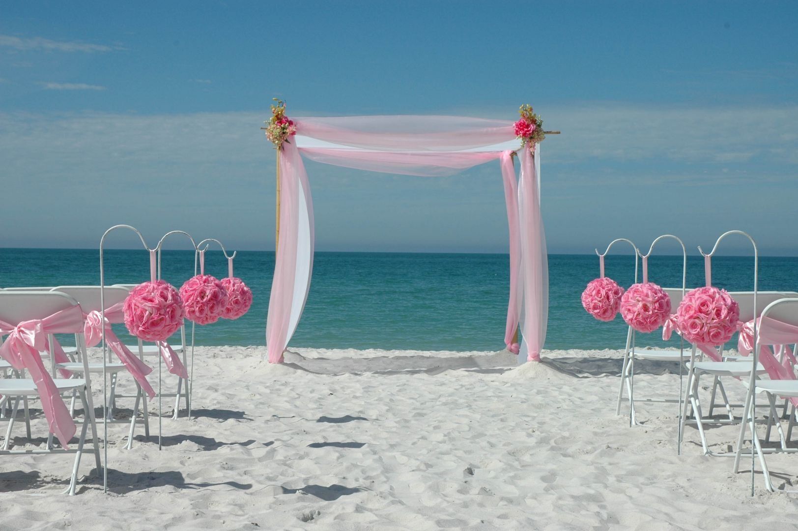 Beach wedding setup with pink fabric and floral accents on chairs and archway.