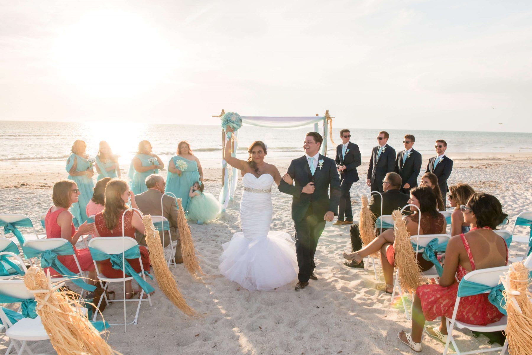 Wedding couple walking down sandy beach aisle; bridesmaids, groomsmen, and guests watch under a sunny sky.