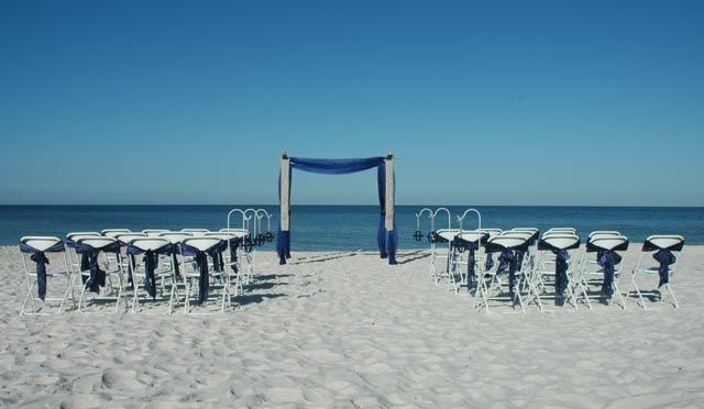 Beach wedding setup with chairs facing a draped arch, ocean backdrop.