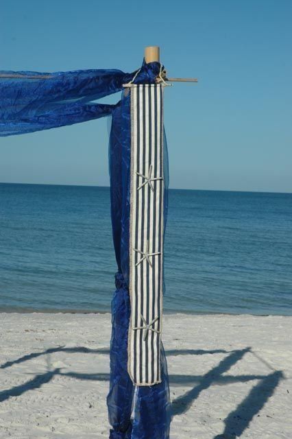 Blue fabric drapes on a striped pillar on a beach, ocean in background.