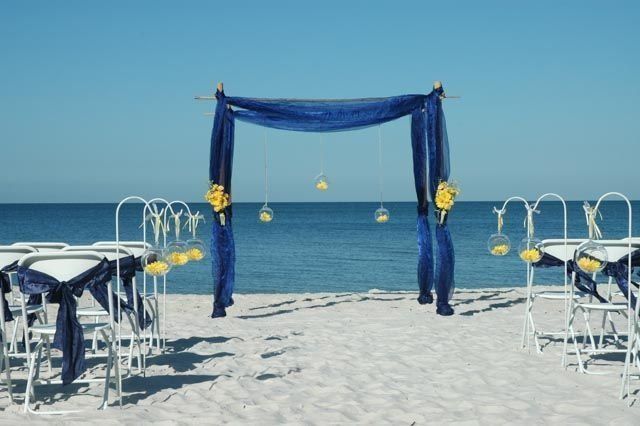 Beach wedding setup: blue draped arch, white chairs with blue sashes, yellow flowers, ocean backdrop.