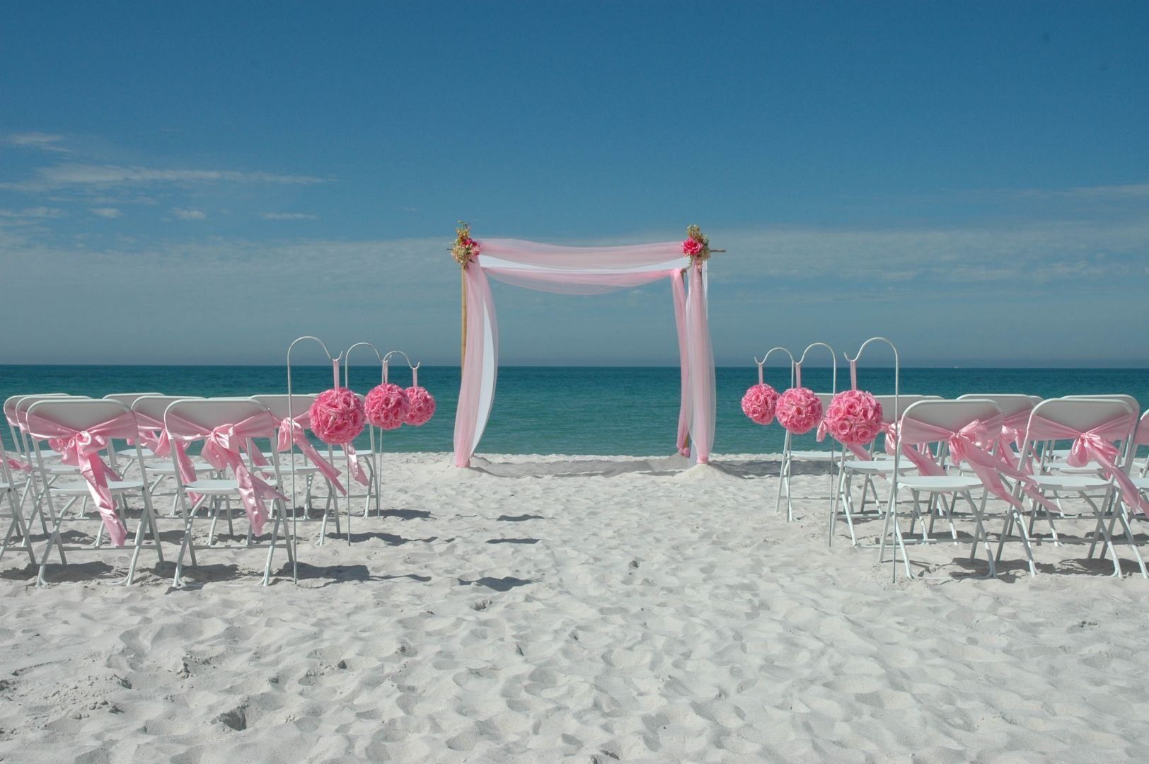 Beach wedding setup with pink decorations and arch; rows of white chairs face the ocean.