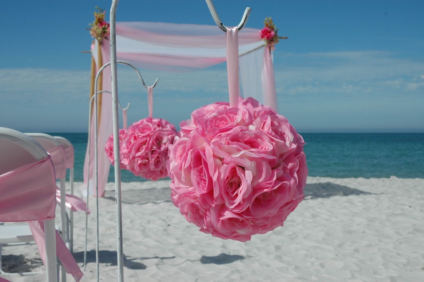 Pink floral spheres hanging on a beach wedding arch with a blue sky and ocean in the background.