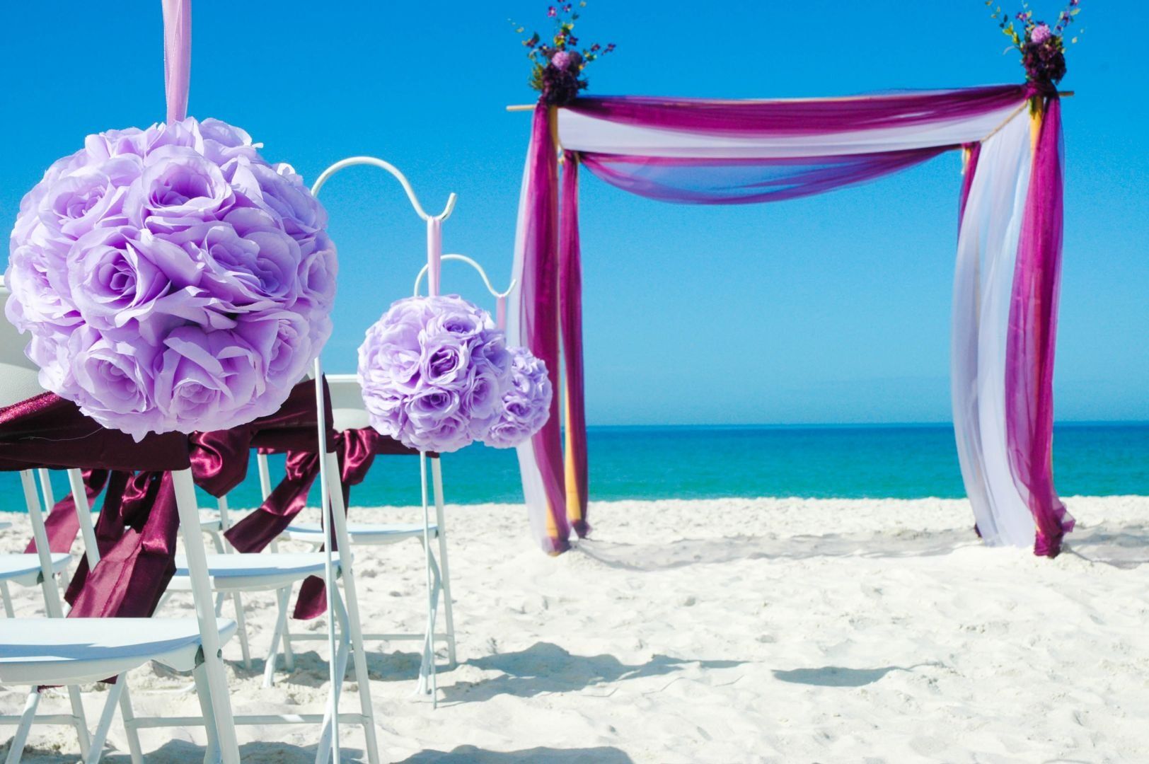Beach wedding setup with purple and white decorations: arch, chairs, and floral spheres.