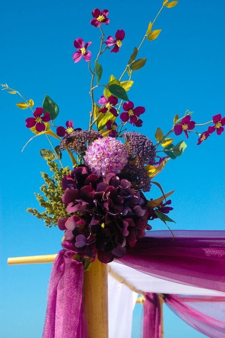 Purple floral arrangement on a wedding arch with draped fabric against a blue sky.