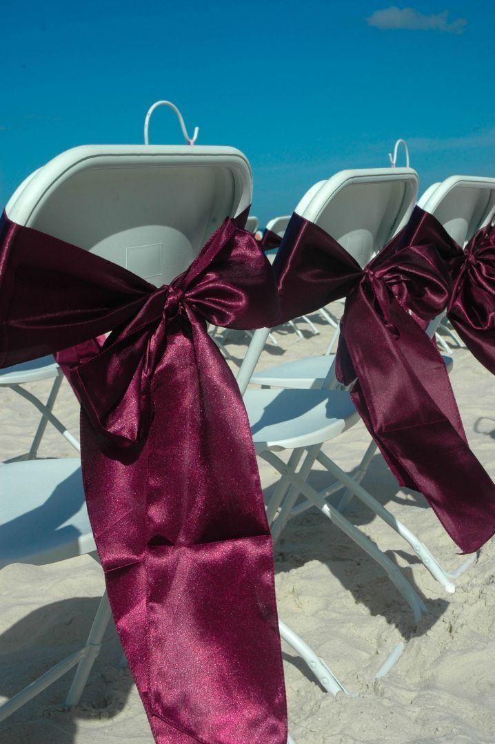 White folding chairs with maroon bows set on a beach with a blue sky.