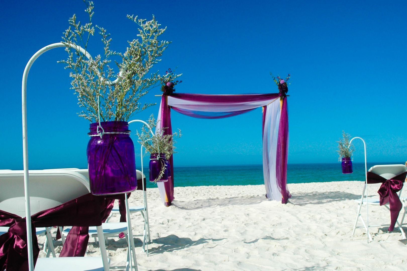 Beach wedding setup with purple decorations against blue sky and ocean.