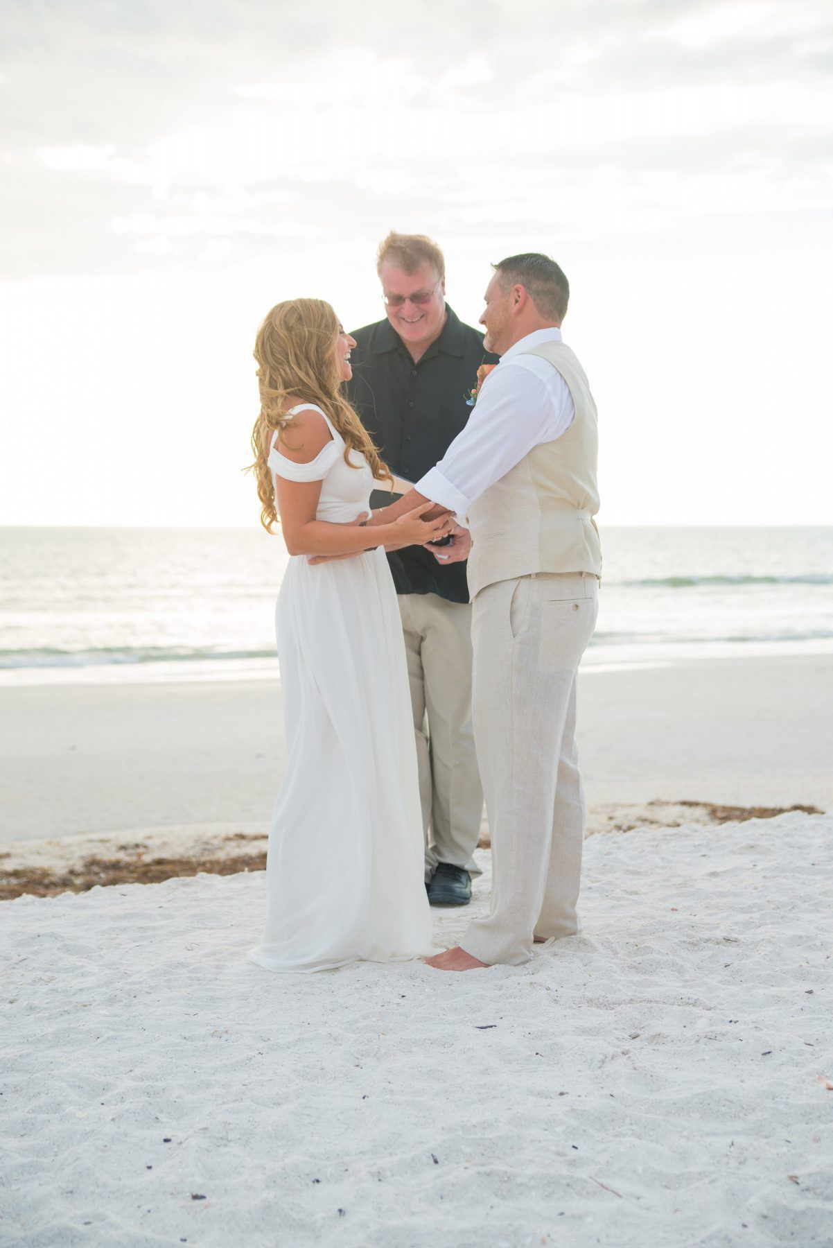 Couple exchanging vows on a beach, officiant present. Bride in white dress, groom in vest. Sunset.