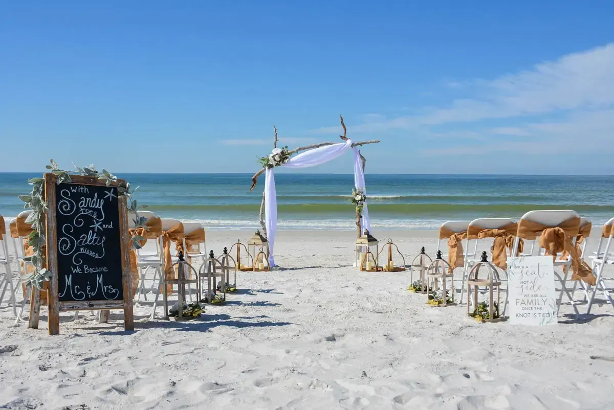 Beach wedding ceremony setup with white chairs, arch, and signage; blue ocean in background.