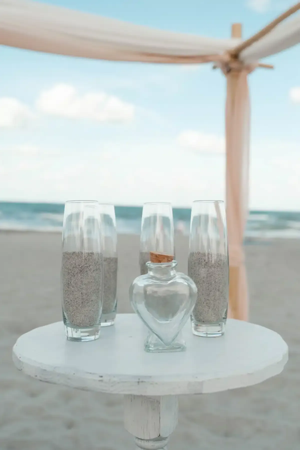 Sand ceremony setup on a beach: white table, clear vases with sand, heart-shaped bottle, arch, ocean backdrop.