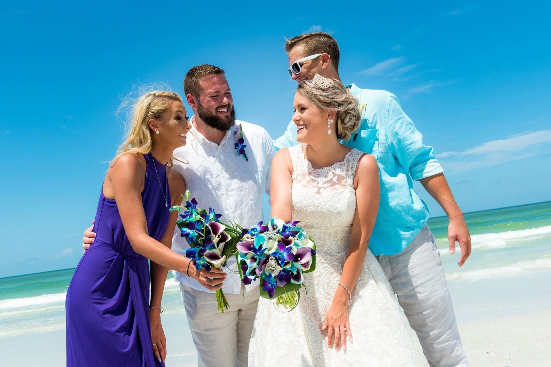 Wedding party on a sunny beach; bride holding flowers smiles, others laugh. Turquoise water and sky.