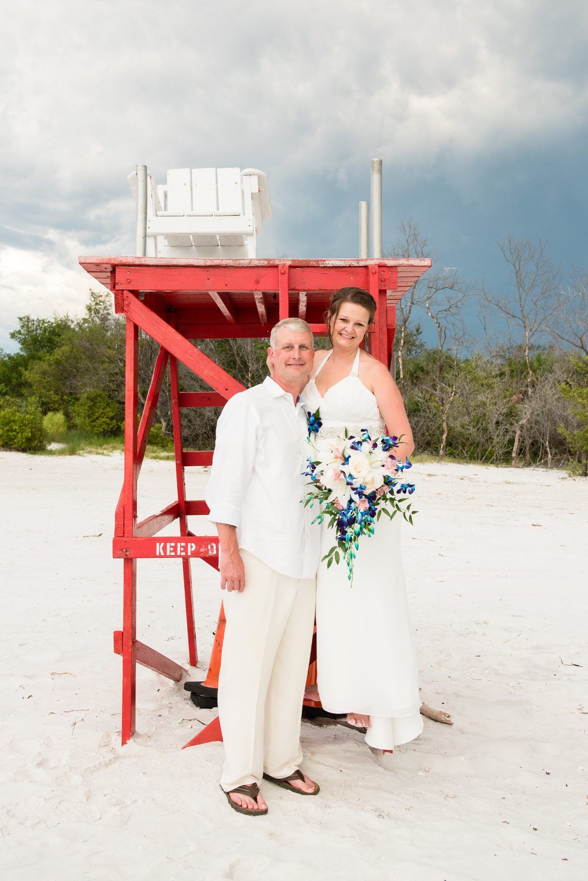 Couple in wedding attire posing by a red lifeguard stand on a beach, holding a bouquet. Cloudy sky.