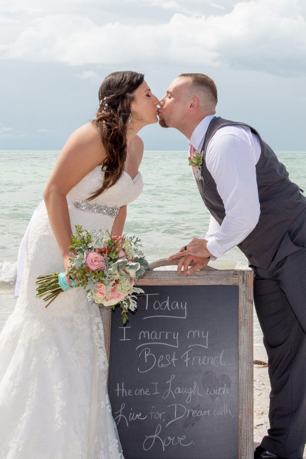 Bride and groom kissing, by the ocean. Bride holds bouquet, wears a white dress. Sign says, 