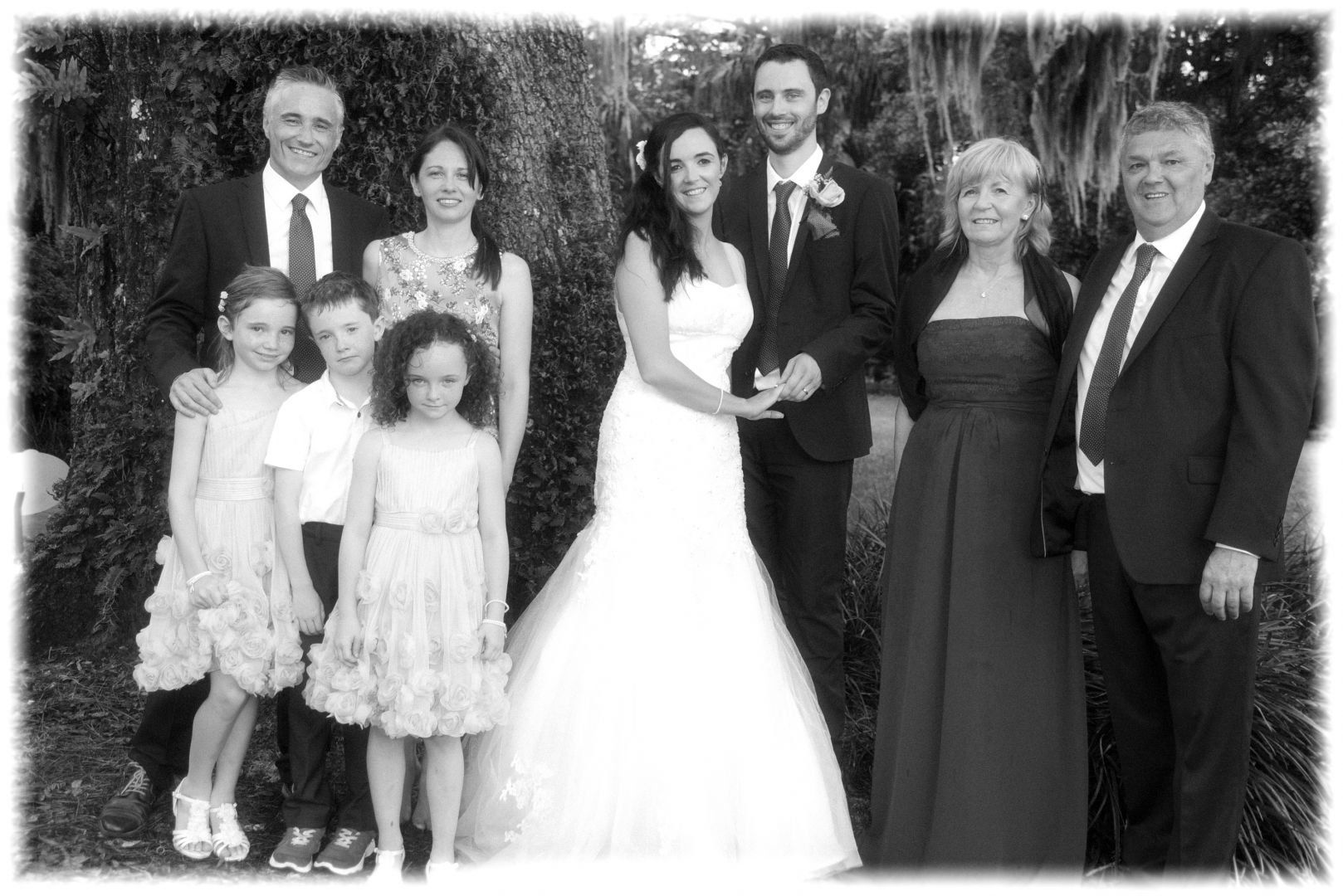 Wedding group photo in black and white: bride and groom with family in front of greenery, smiling.