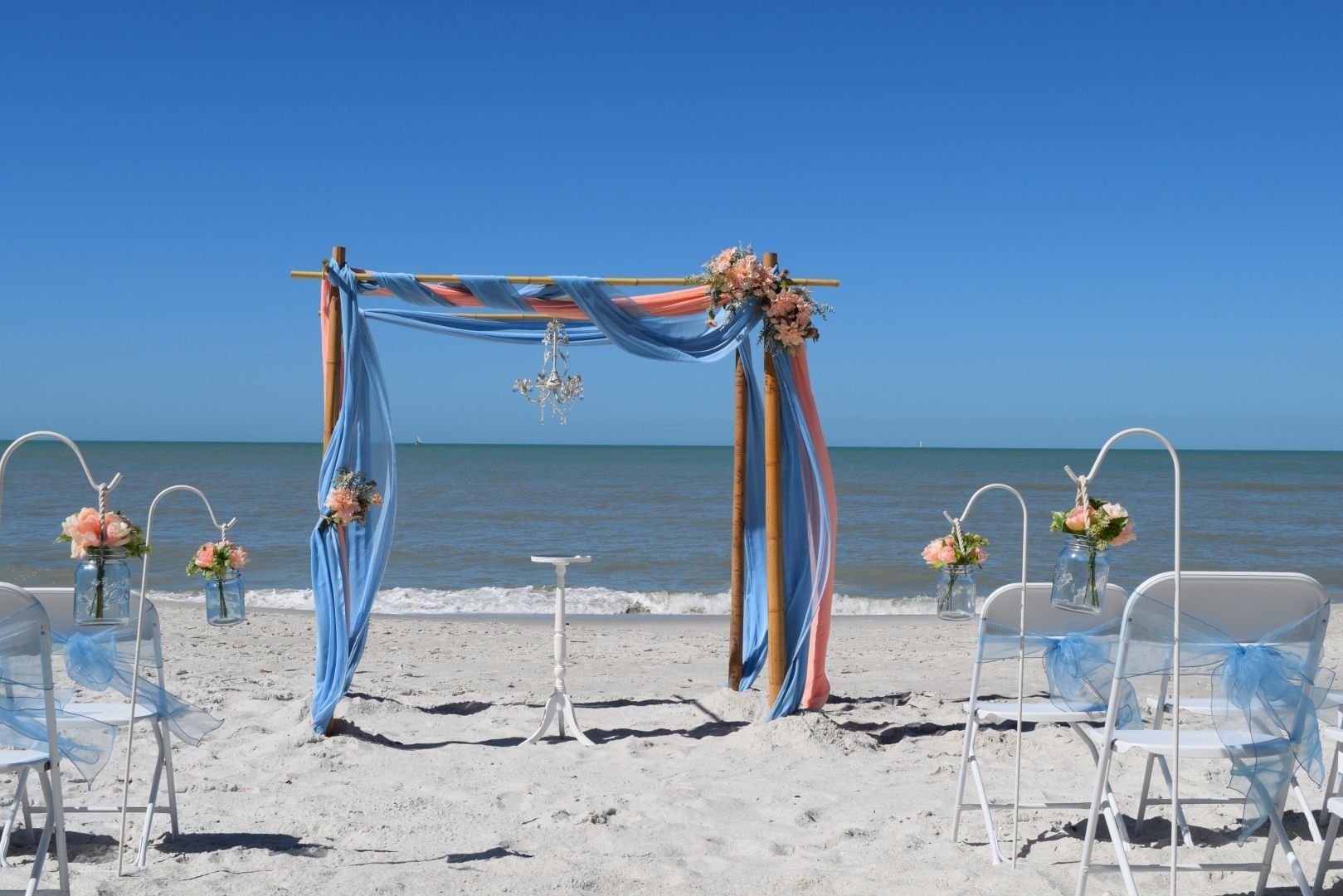 Beach wedding setup with draped arch, chairs, and ocean backdrop.