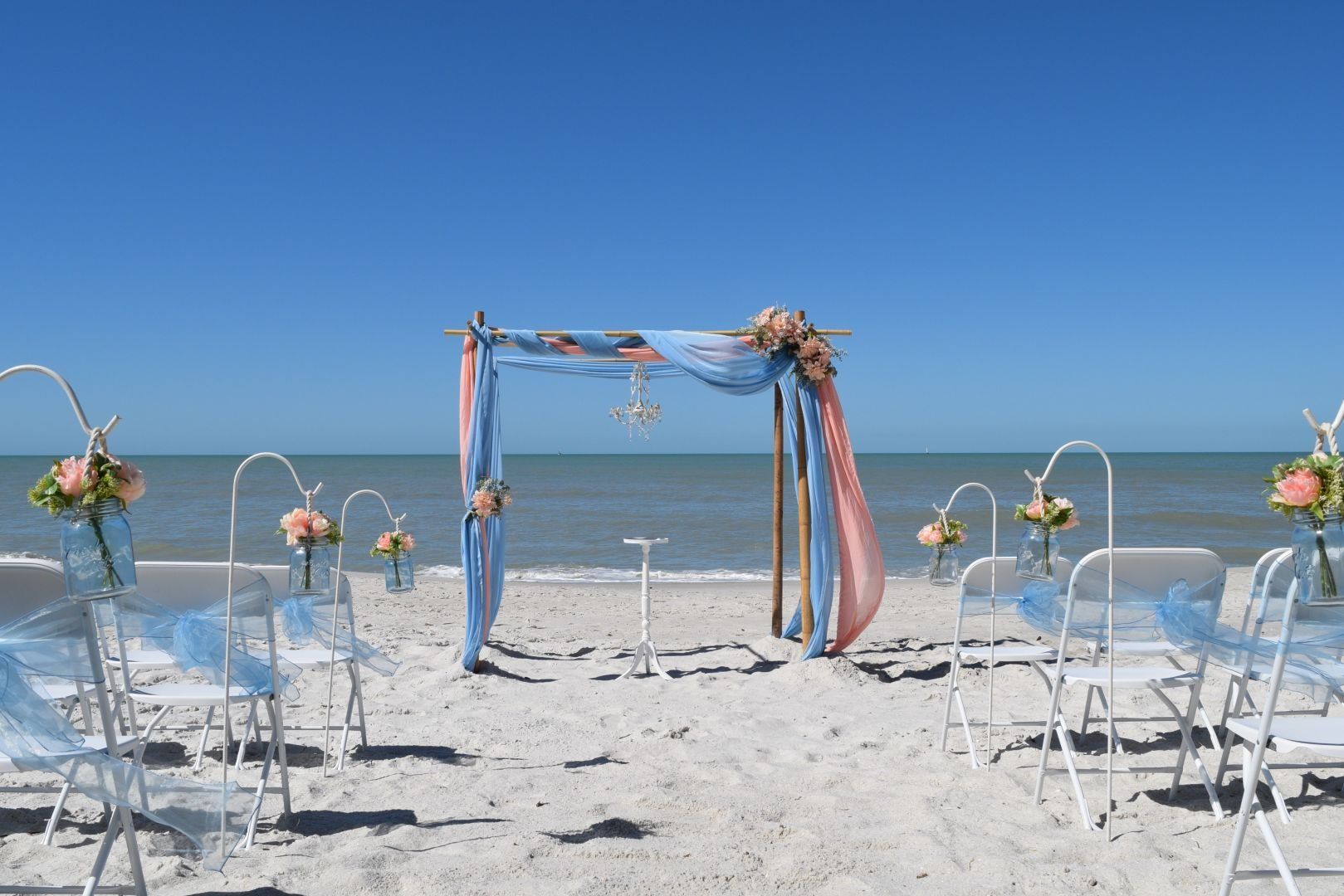 Beach wedding setup: arch with blue and pink fabric, white chairs, ocean and clear sky.