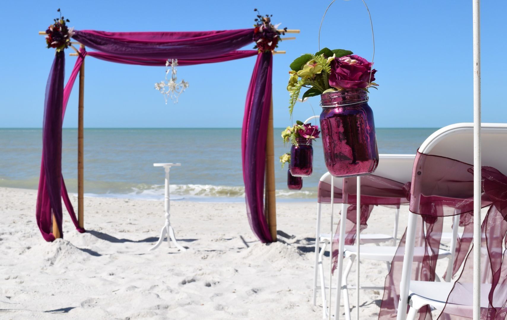 Beach wedding setup with purple draped canopy, floral accents, and chairs.