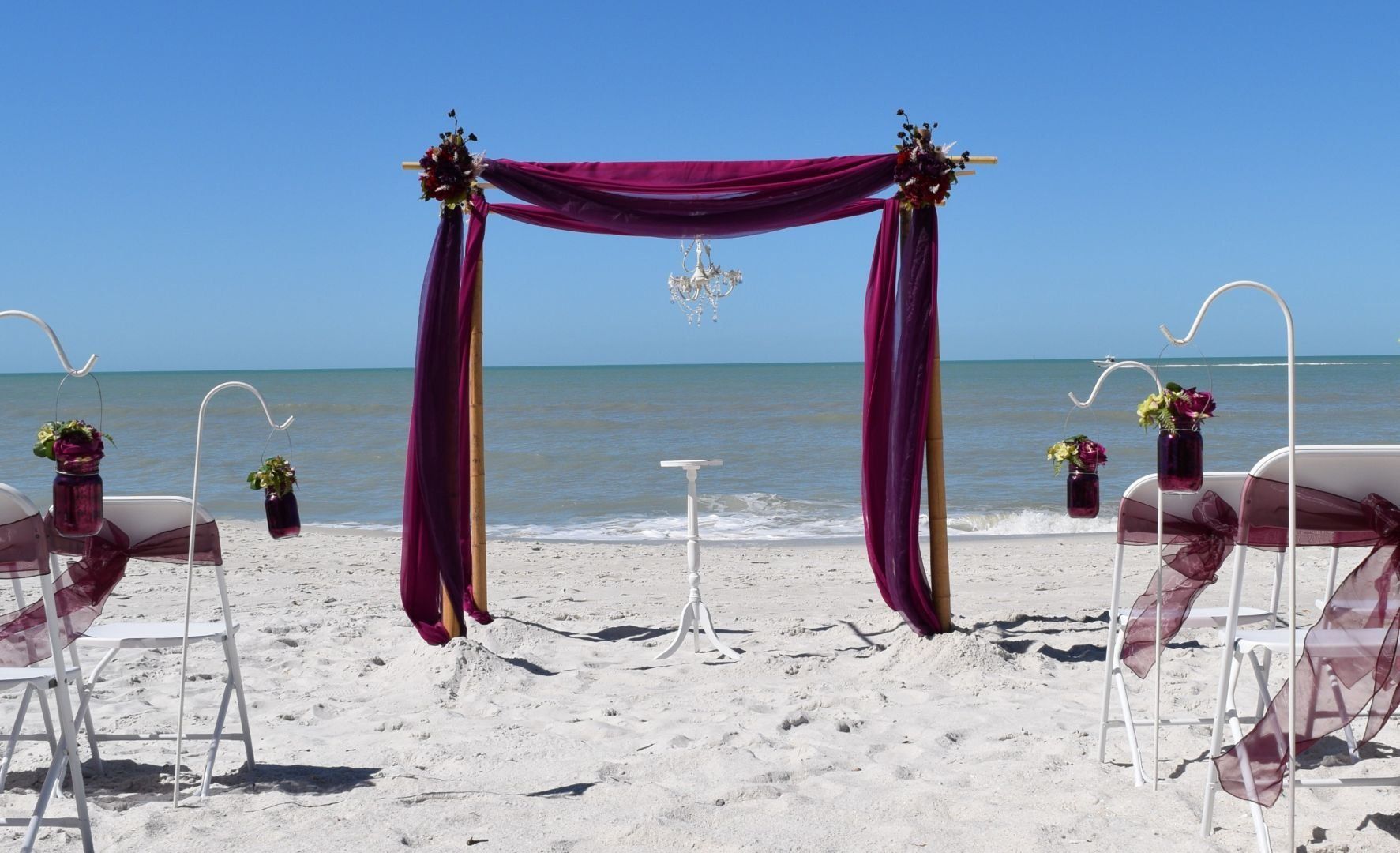 Beach wedding setup with purple draped archway and chairs facing the ocean.