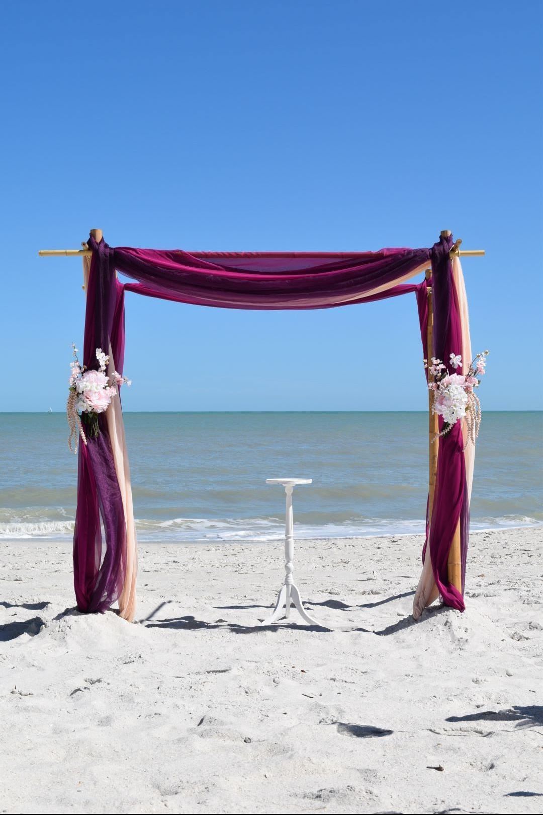 Beach wedding arch with purple and gold fabric, white sand, and blue sky.