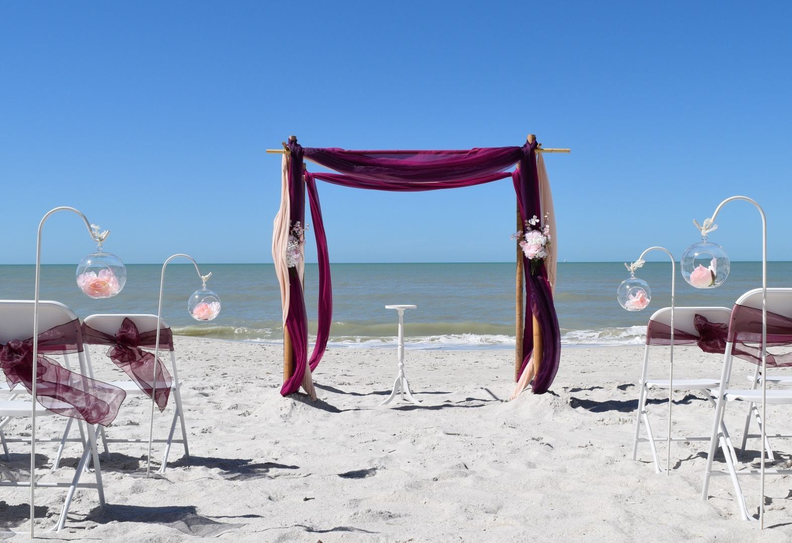 Beach wedding setup: Purple draped arch, chairs, and decorations on white sand, with ocean and blue sky backdrop.