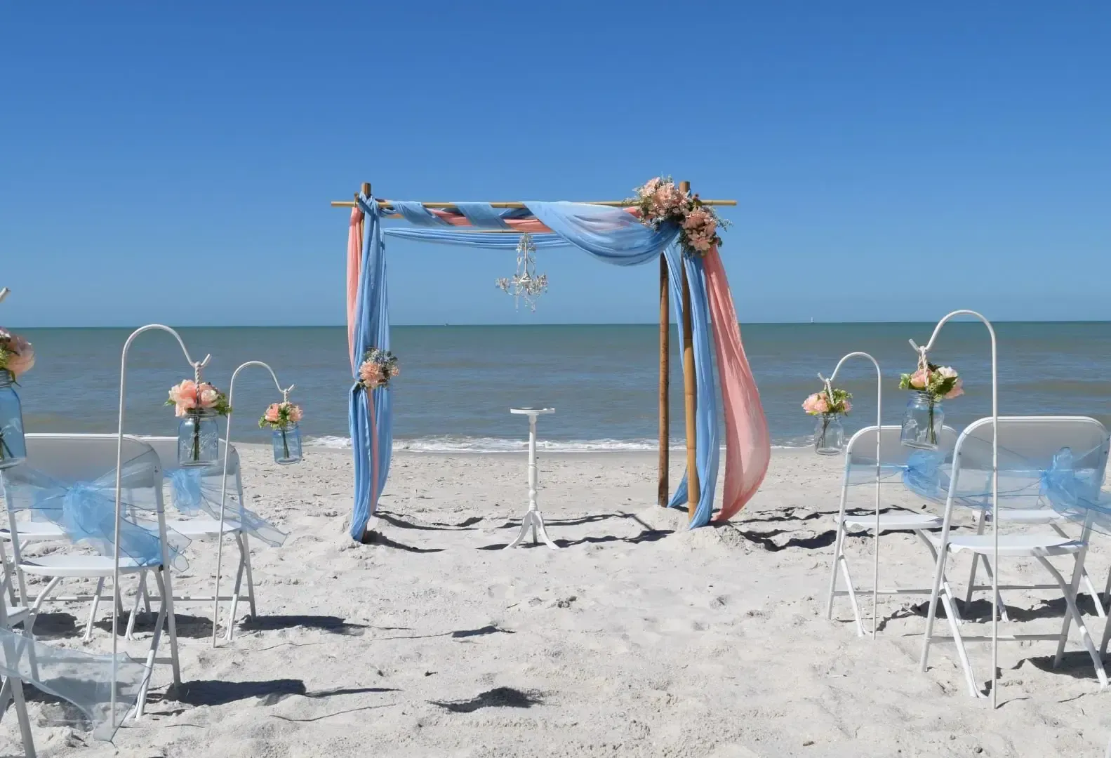 Beach wedding setup with arch draped in peach and blue fabric, chairs, and ocean backdrop.