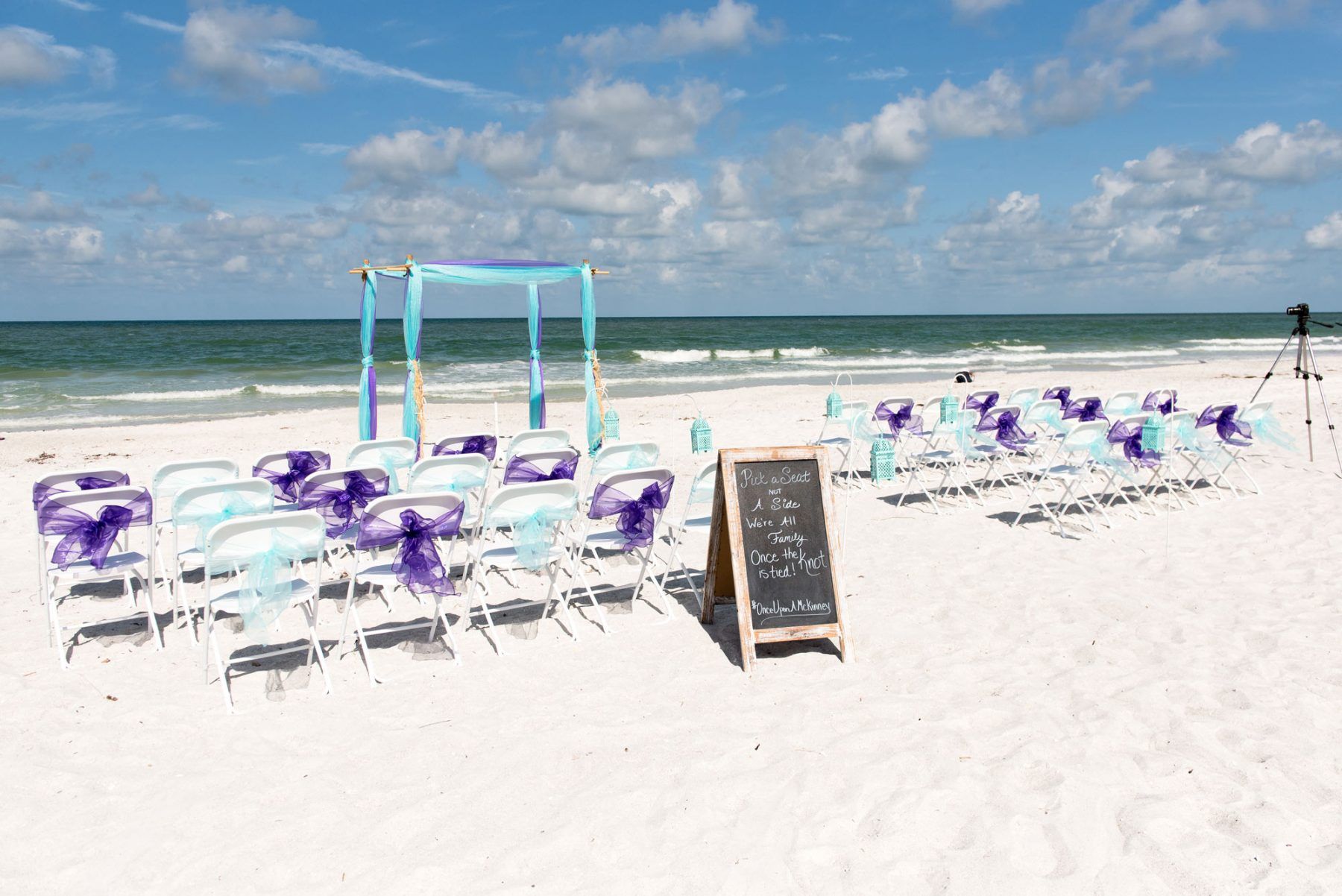 Beach wedding setup with chairs, arch, and sign on a sunny day.