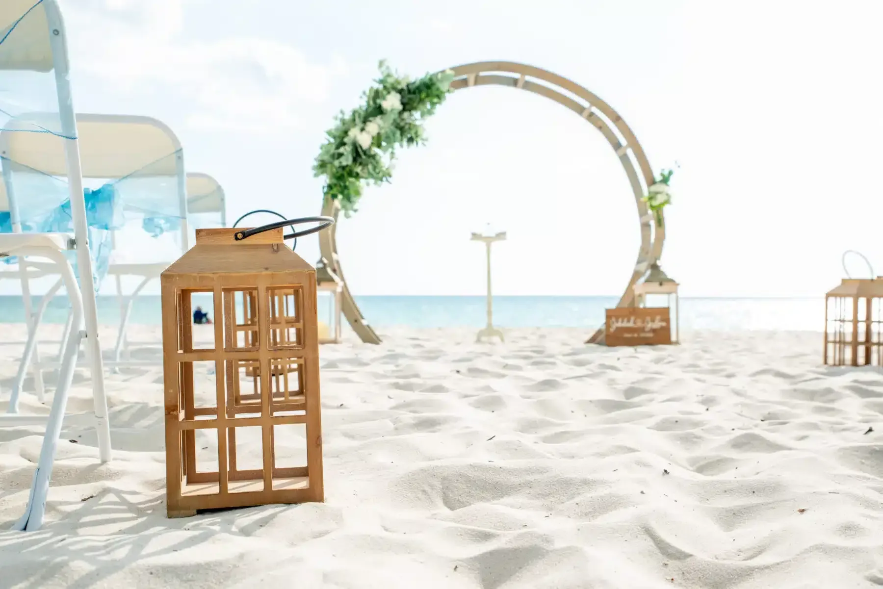 Beach wedding ceremony setup with a floral arch, lanterns, and chairs on white sand.
