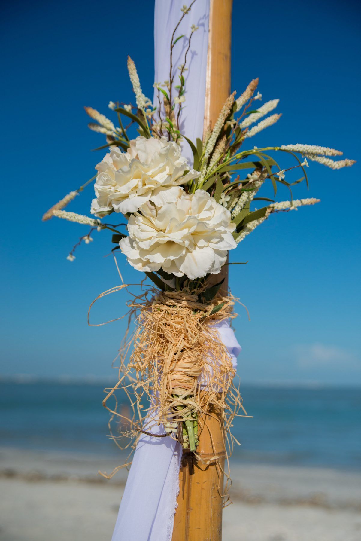 Beach wedding decoration with white flowers, straw, and fabric on a bamboo pole against a blue sky and ocean.