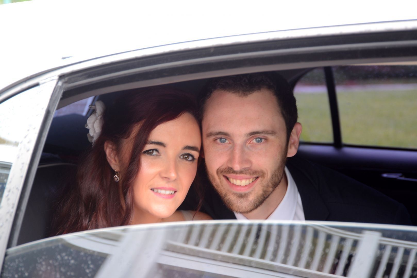 Newly married couple smiling in a car, woman with flower in hair, man in suit.