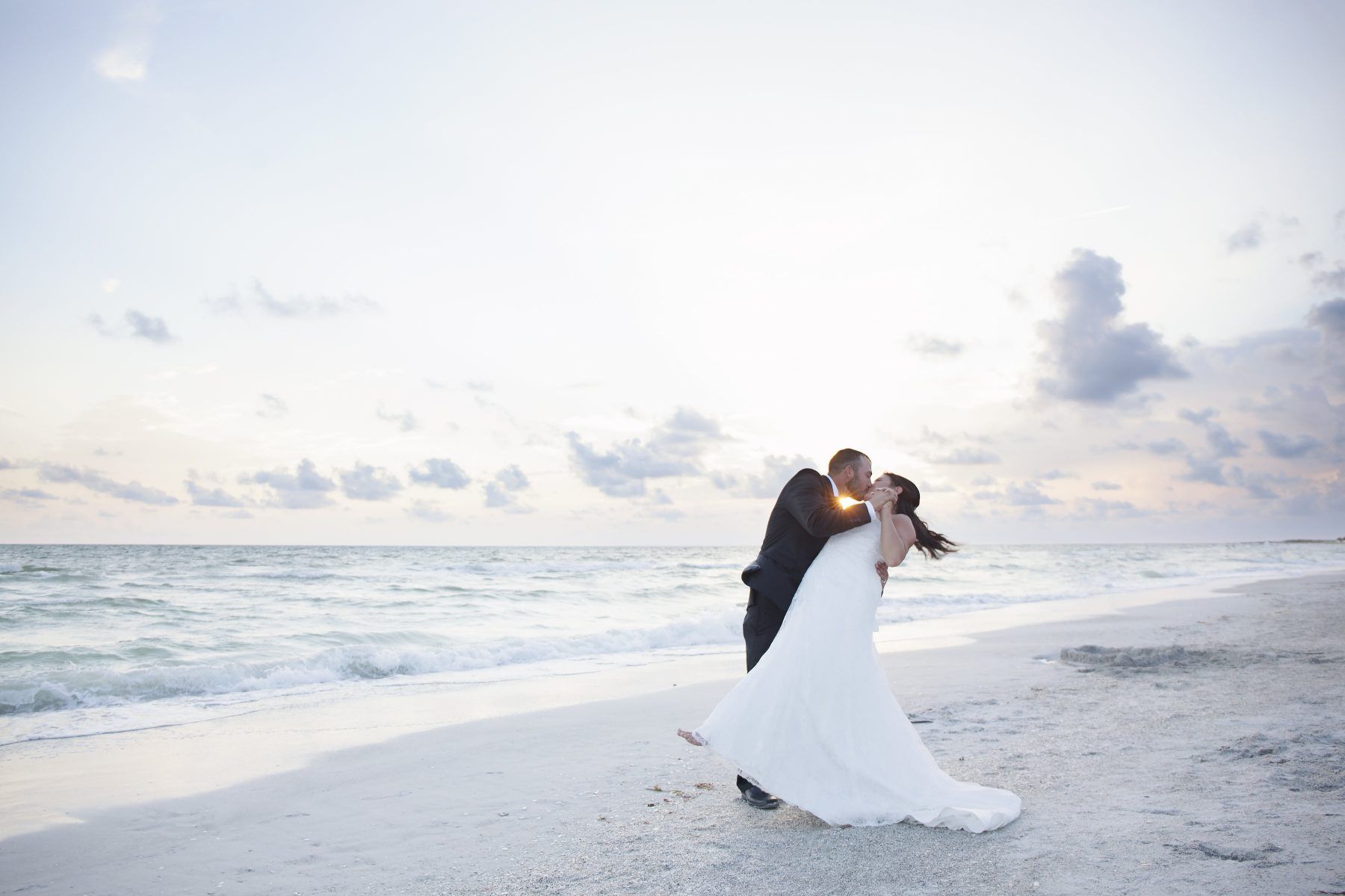 Couple kissing on a beach at sunset; bride in white gown, groom in suit.
