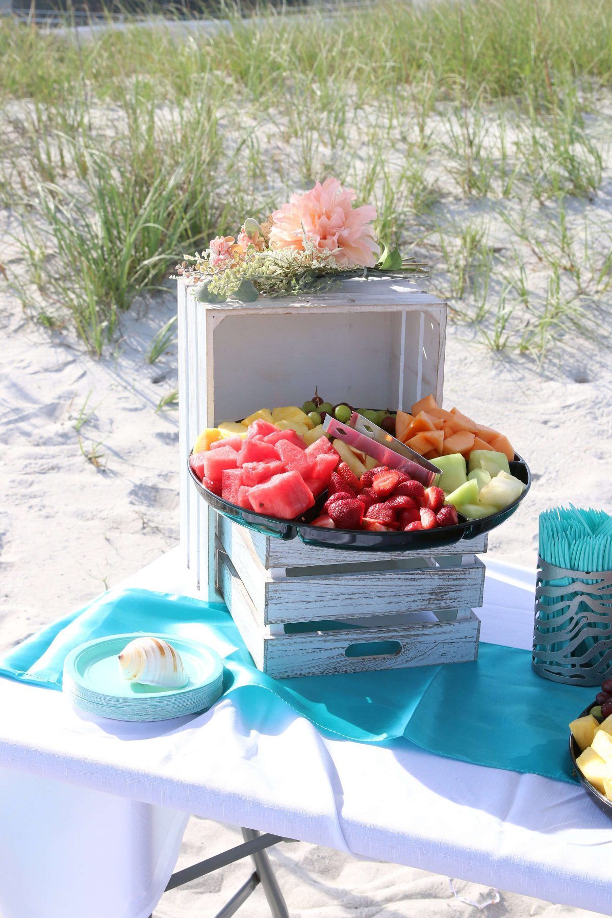 Fruit platter on a beach table, flowers on a crate. Teal accents on white sand background.