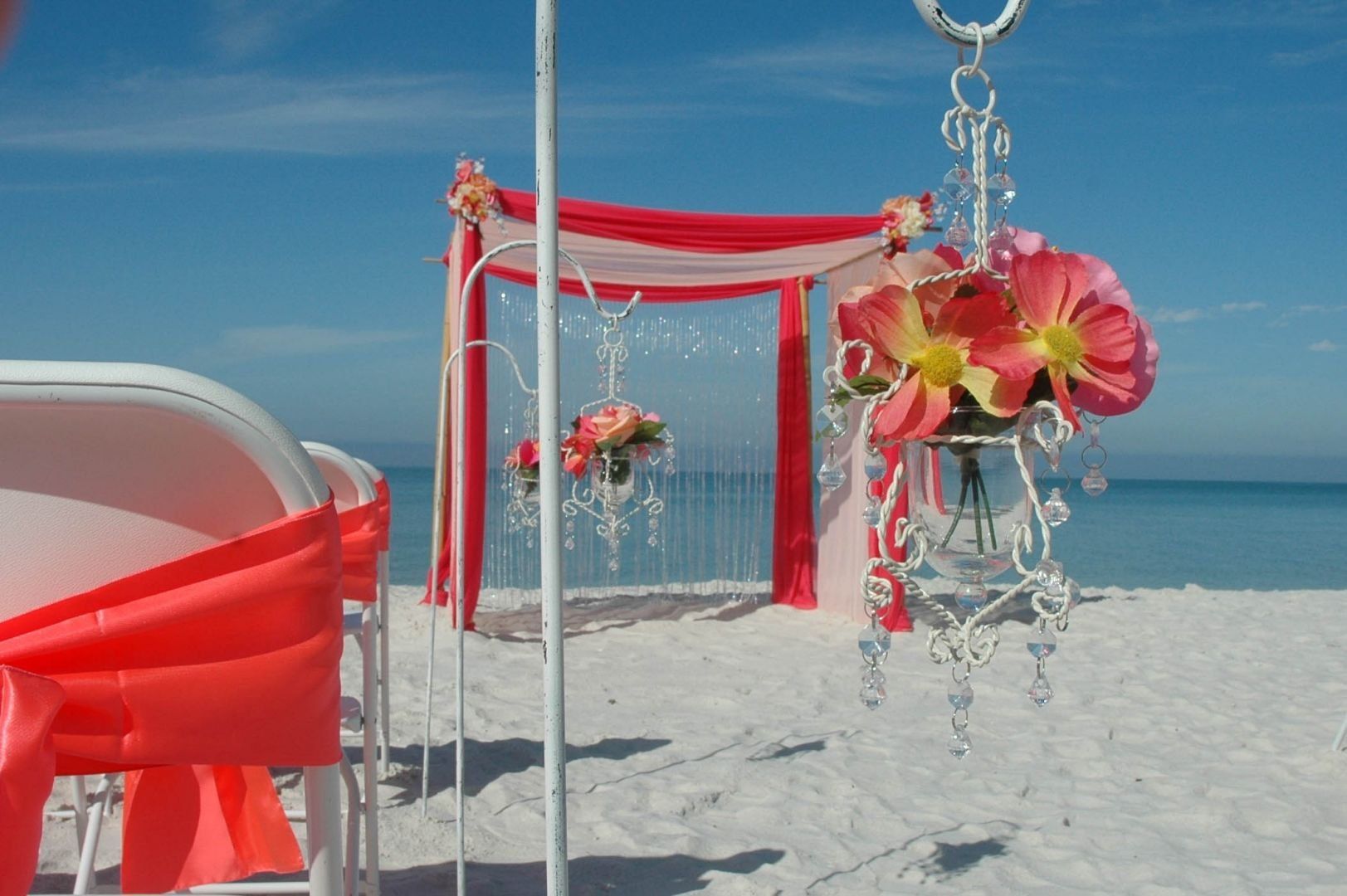 Beach wedding setup with a coral and white archway and floral chandelier decorations.