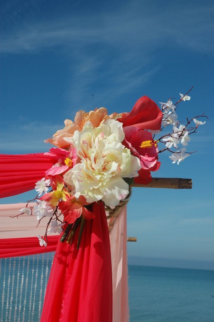 Floral arch with pink and coral fabric, against a bright blue sky and ocean.