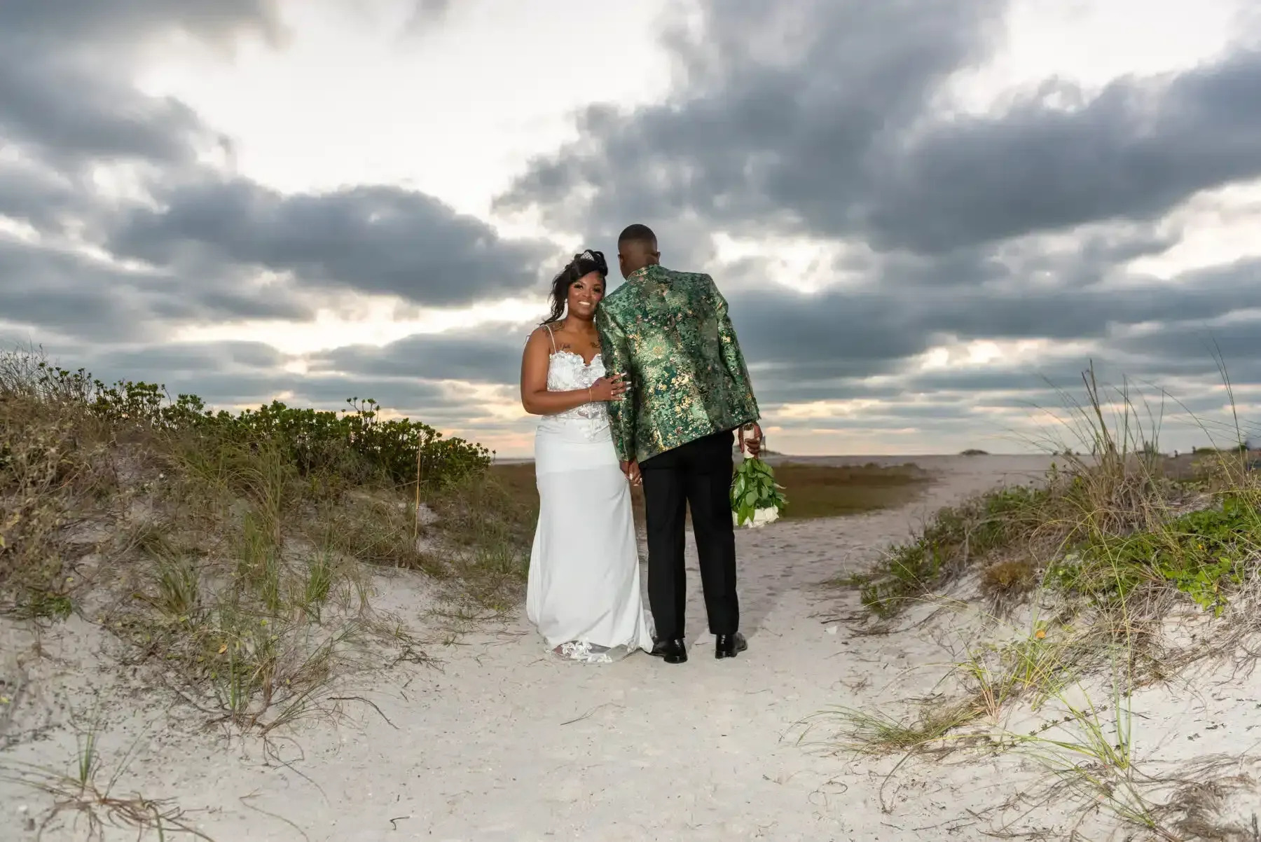 Couple on a sandy path, holding hands, beneath a cloudy sky at the beach. The groom's jacket is patterned.