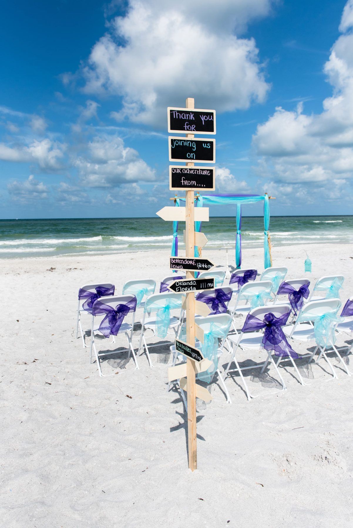 Beach wedding setup with chairs, signpost, and ocean view; blue and purple accents.